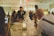A couple, possibly newlyweds, is cutting a cake with a professional photographer taking pictures of the moment. The setting appears to be a warmly lit reception area with other guests in the background. A smartphone is visible in the foreground, capturing the event.