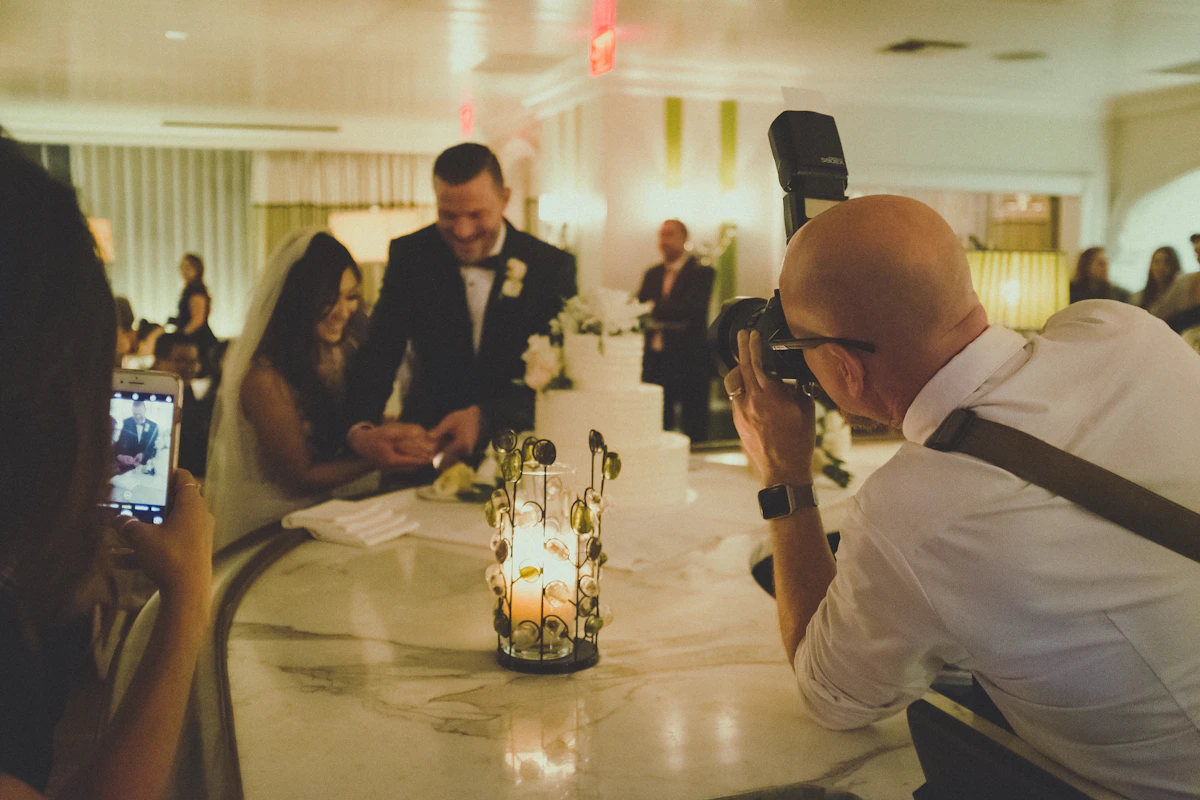 A photographer shooting a couple at a cake table while a guest films on a phone
