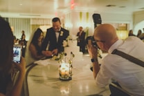 A couple, possibly newlyweds, is cutting a cake with a professional photographer taking pictures of the moment. The setting appears to be a warmly lit reception area with other guests in the background. A smartphone is visible in the foreground, capturing the event.
