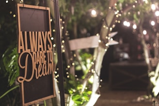 A warmly lit sign with the words 'Always dare to dream' hangs among lush greenery and twinkling fairy lights, evoking a cozy and inviting atmosphere.