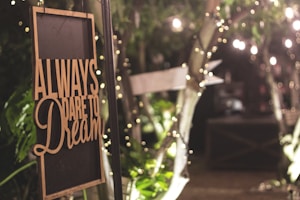 A warmly lit sign with the words 'Always dare to dream' hangs among lush greenery and twinkling fairy lights, evoking a cozy and inviting atmosphere.