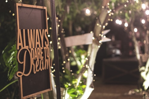 A warmly lit sign with the words 'Always dare to dream' hangs among lush greenery and twinkling fairy lights, evoking a cozy and inviting atmosphere.