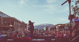 A large crowd gathers in an urban setting, watching a live music performance with mountains visible in the background. The scene includes a band performing on an outdoor stage, with musicians silhouetted against the early evening sky. Brick buildings line the sides of the street, contributing to the lively atmosphere.