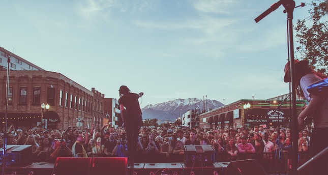 A large crowd gathers in an urban setting, watching a live music performance with mountains visible in the background. The scene includes a band performing on an outdoor stage, with musicians silhouetted against the early evening sky. Brick buildings line the sides of the street, contributing to the lively atmosphere.