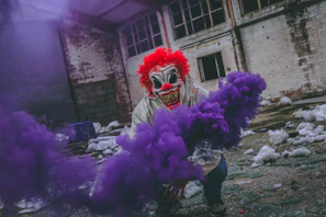 A grinning clown with cracked makeup standing in a foggy, abandoned carnival tent.