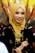 A woman is wearing a golden hijab adorned with an elegant tiara and intricate jewelry. Her attire features gold embellishments that complement the detailed accessories, and she is smiling warmly.