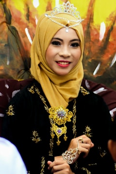 A woman is wearing a golden hijab adorned with an elegant tiara and intricate jewelry. Her attire features gold embellishments that complement the detailed accessories, and she is smiling warmly.