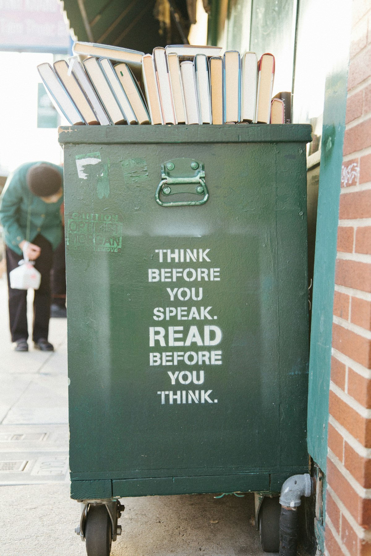 Books on a green cart next to a brick wall.  White text in stencil says "THINK BEFORE YOU SPEAK.  READ BEFORE YOU THINK."  The word READ is twice as large.