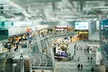 A busy airport terminal with numerous travelers walking through a large open area. There are multiple levels with escalators leading down to the main concourse. Various shops and eateries line the sides, and the space is filled with bright lighting and modern architecture.