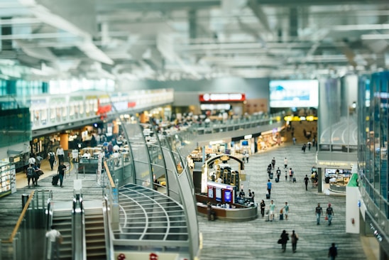 A busy airport terminal with numerous travelers walking through a large open area. There are multiple levels with escalators leading down to the main concourse. Various shops and eateries line the sides, and the space is filled with bright lighting and modern architecture.