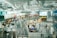 A busy airport terminal with numerous travelers walking through a large open area. There are multiple levels with escalators leading down to the main concourse. Various shops and eateries line the sides, and the space is filled with bright lighting and modern architecture.