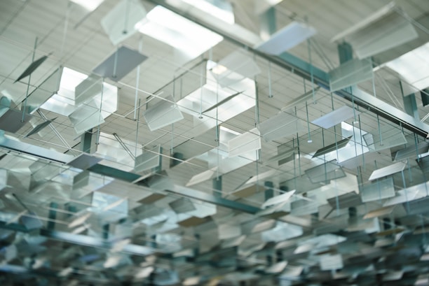 A complex arrangement of metallic sheets suspended from the ceiling. The sheets are geometrically shaped, reflecting light, and are mounted with thin rods. The ceiling has a modern architectural design with alternating patterns and a somewhat transparent appearance.