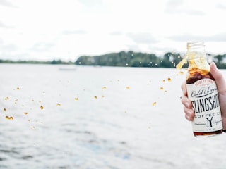A hand is holding a bottle of cold brew coffee labeled 'Slingshot', near a body of water. Liquid is splashing out of the open bottle, with droplets suspended in the air. In the background, there is a blurred view of trees and an overcast sky.