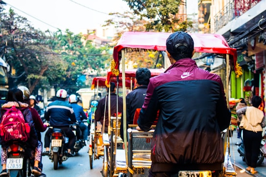 A busy street scene featuring several people riding bicycles and motorbikes. The motorbikes are in motion, weaving through traffic. The environment indicates a lively urban atmosphere with trees and buildings in the background. People are wearing casual clothing, and the presence of helmets suggests a focus on safety. Various shades and colors are evident in the clothing and vehicles, highlighting a vibrant street life.