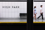 A subway platform with tiled walls features the words 'HIGH PARK' in bold black letters. A metal bench is visible beneath the sign, and two people are captured in motion blur as they walk past. The platform edge is marked with a yellow strip for safety.