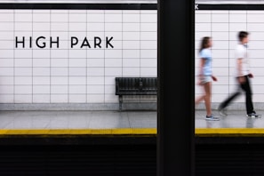 A subway platform with tiled walls features the words 'HIGH PARK' in bold black letters. A metal bench is visible beneath the sign, and two people are captured in motion blur as they walk past. The platform edge is marked with a yellow strip for safety.