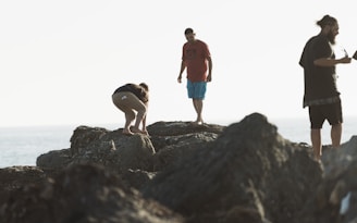 Three people are standing on rugged rocks near the shoreline. One person is crouched down, examining something on the ground, while another stands with their feet together, gazing down. The third person is holding an object, possibly a coffee cup, and looking towards the distance. The scene is softly lit by natural light, creating a tranquil and adventurous atmosphere.