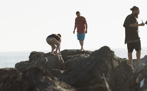 Three people are standing on rugged rocks near the shoreline. One person is crouched down, examining something on the ground, while another stands with their feet together, gazing down. The third person is holding an object, possibly a coffee cup, and looking towards the distance. The scene is softly lit by natural light, creating a tranquil and adventurous atmosphere.