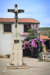 A stone statue of Jesus on a crucifix stands prominently in front of a small building with a tiled roof. To the side, purple flowering bougainvillea adds color to the scene. A green street lamp is also visible nearby.
