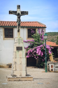 A stone statue of Jesus on a crucifix stands prominently in front of a small building with a tiled roof. To the side, purple flowering bougainvillea adds color to the scene. A green street lamp is also visible nearby.