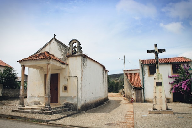 A small, rustic chapel with a bell on top is situated on a cobblestone street, accompanied by a cross monument on the right. The buildings have red-tiled roofs, and there is a flowering bush adding a touch of color.