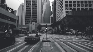 A monochrome image of a bustling urban street surrounded by tall skyscrapers. Cars and pedestrians are visible, creating a sense of busy city life. The grid-like road markings and overhead wires add to the metropolitan atmosphere.