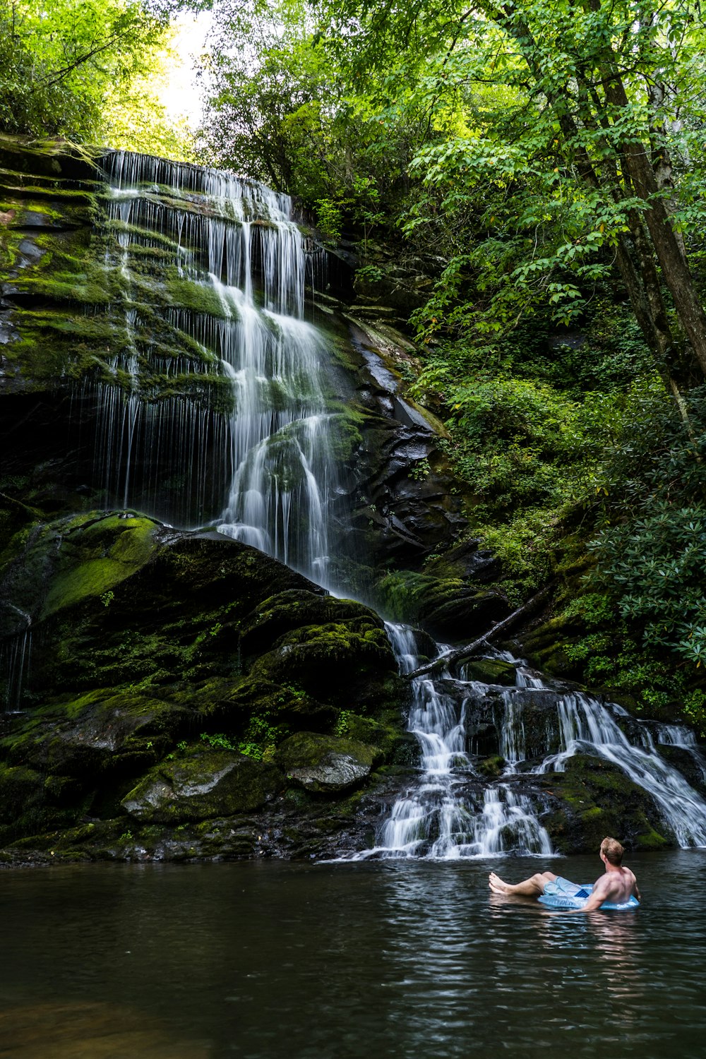 man riding blue inflatable ring in body of water while looking up watching the waterfalls at daytime