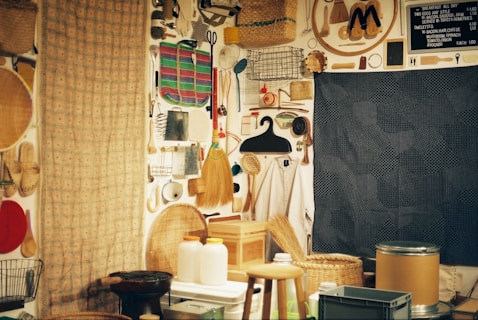 A room with a wall displaying a diverse assortment of household items and tools arranged for decorative or storage purposes. The collection includes brooms, baskets, hand tools, and various kitchen utensils. In the foreground, there are stools, containers, and jars. The interior has a cozy and eclectic feel with a menu board listing breakfast items.