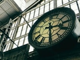 A large, vintage clock with black Roman numerals is mounted on a metal structure inside an industrial building. The clock face is slightly aged and surrounded by tall, paneled windows that allow light to filter through. Metal beams and a corrugated roof add to the industrial atmosphere.