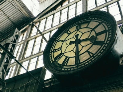 A large, vintage clock with black Roman numerals is mounted on a metal structure inside an industrial building. The clock face is slightly aged and surrounded by tall, paneled windows that allow light to filter through. Metal beams and a corrugated roof add to the industrial atmosphere.