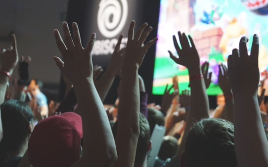 A crowd of people with hands raised in an enthusiastic gesture, standing in front of a large screen displaying colorful graphics. The setting suggests a lively event or gathering. Some individuals wear wristbands, hinting at an organized occasion.