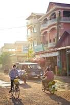 A bustling street scene in a tropical location featuring a few people riding bicycles and a motorbike on a dirt road. The background includes a row of shops and buildings with signs in a Southeast Asian script. The sunlight casts a warm glow, creating shadows on the street.