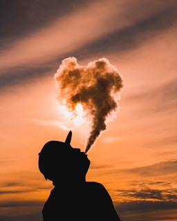 A person is silhouetted against a vibrant sunset sky, exhaling a cloud of vapor that is illuminated by the sun, creating a heart-shaped formation in the air. The warm colors of the sunset create a dramatic and artistic backdrop.