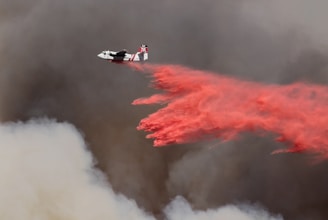 A firefighting aircraft releases a large stream of red fire retardant over a smoky, dark sky. The contrast between the bright red liquid and the surrounding gray and brown smoke is striking, emphasizing the urgency of the situation.