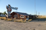 A rustic log cabin-like building with a vintage sign advertising 'Tumble Inn' and 'Sizzlin Steaks.' The building has a worn appearance and is surrounded by a gravel lot and overgrown grass. A large metal tower stands nearby, and an RV is parked to the left of the building.