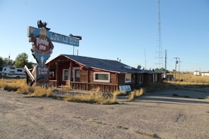 A rustic log cabin-like building with a vintage sign advertising 'Tumble Inn' and 'Sizzlin Steaks.' The building has a worn appearance and is surrounded by a gravel lot and overgrown grass. A large metal tower stands nearby, and an RV is parked to the left of the building.