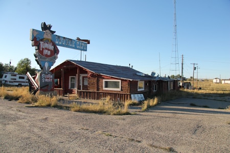 A rustic log cabin-like building with a vintage sign advertising 'Tumble Inn' and 'Sizzlin Steaks.' The building has a worn appearance and is surrounded by a gravel lot and overgrown grass. A large metal tower stands nearby, and an RV is parked to the left of the building.
