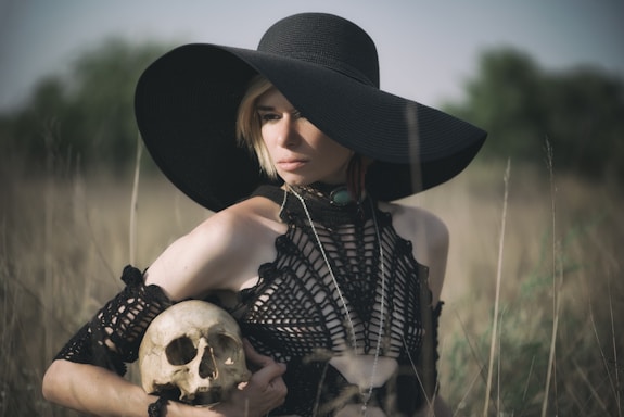 A person wearing a large black hat and a crochet-style black dress is holding a human skull. The background is an open field of tall, dry grass and blurred trees, suggesting an outdoor setting. The composition and attire exude a gothic or mysterious feel.