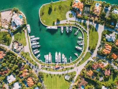 An aerial view of a marina surrounded by luxurious houses with lush green gardens. The marina is filled with several yachts and boats docked in a semicircular pattern within a calm, teal-colored waterway. The surrounding area features organized streets, vibrant greenery, and palm trees adding a tropical atmosphere.