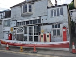 A weathered building with the sign 'Roseland Gig Club' above, displaying vintage style and age. In front, two old-fashioned red and cream Shell petrol pumps stand near a long, red rowboat marked 'Roseland.' Traffic cones are placed around it on the street.