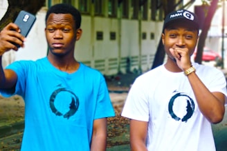 Two individuals are captured outdoors, both wearing t-shirts featuring a circular design. The person on the left, wearing a blue shirt, is holding a smartphone as if taking a selfie. The person on the right, in a white shirt and black cap, is covering their mouth with one hand and appears amused.