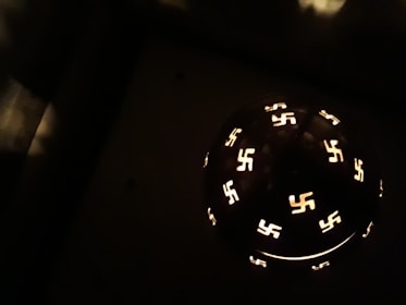 A darkened setting with a spherical object featuring multiple illuminated swastika symbols arranged around its surface, casting a glow in otherwise shadowy surroundings.