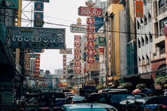 A bustling street scene filled with various signs in Thai and Chinese characters, surrounded by buildings. The area is busy with vehicles like tuk-tuks and cars. The signs are prominently visible, featuring bold colors and intricate designs.