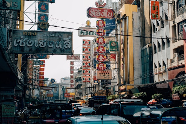 A bustling street scene filled with various signs in Thai and Chinese characters, surrounded by buildings. The area is busy with vehicles like tuk-tuks and cars. The signs are prominently visible, featuring bold colors and intricate designs.