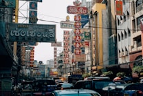 A bustling street scene filled with various signs in Thai and Chinese characters, surrounded by buildings. The area is busy with vehicles like tuk-tuks and cars. The signs are prominently visible, featuring bold colors and intricate designs.