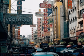 A bustling street scene filled with various signs in Thai and Chinese characters, surrounded by buildings. The area is busy with vehicles like tuk-tuks and cars. The signs are prominently visible, featuring bold colors and intricate designs.