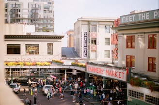 An outdoor farmers market bustling with people, surrounded by city buildings. The market has a prominent red sign. Floral arrangements decorate the roofline, and the scene is lively with vendors and visitors.