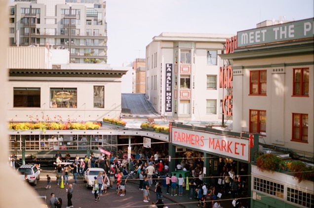An outdoor farmers market bustling with people, surrounded by city buildings. The market has a prominent red sign. Floral arrangements decorate the roofline, and the scene is lively with vendors and visitors.