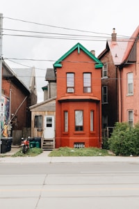 A bright red brick townhouse with a green trim stands between two other homes. It features two arched windows on the upper floor and two vertical windows on the ground floor. The door is adjacent to the house, with a small wooden porch covered with a tan exterior leading to it. Electrical wires are overhead, and a motorcycle, trash cans, and recycling bins are on the left side of the house near the porch. The front yard is sparse with greenery, and the pavement and street occupy the foreground.
