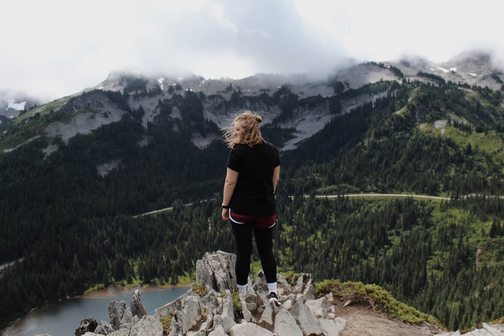 woman standing on rock cliff overlooking trees and body of water with mountains at distance during daytime